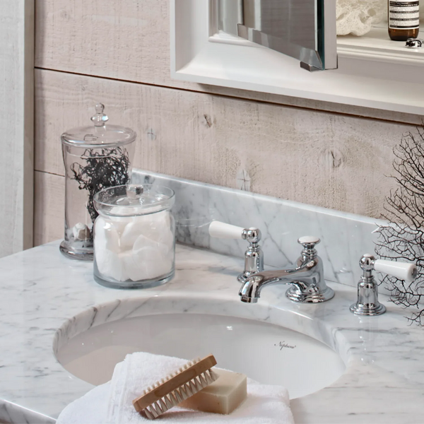 Decorative glass canisters on a white marble bathroom vanity sink. The sink is styled with a wooden scrub brush, a bar of soap on a white towel.