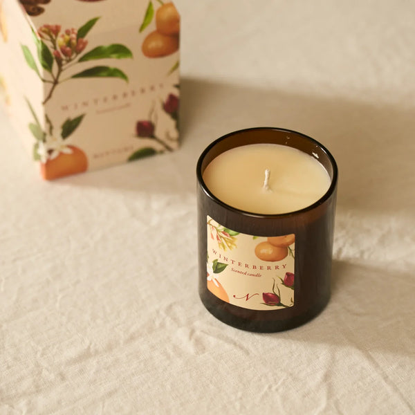 An amber glass jar candle with a cream-colored wick and label, placed on a textured cream fabric surface with its matching illustrated box blurred in the background.