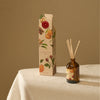 A reed diffuser bottle with amber glass and natural reeds sits beside its illustrated product box on a light fabric surface. The packaging and bottle label show winterberry, citrus, cinnamon, and floral illustrations, with the brand name Nature's Own visible.
