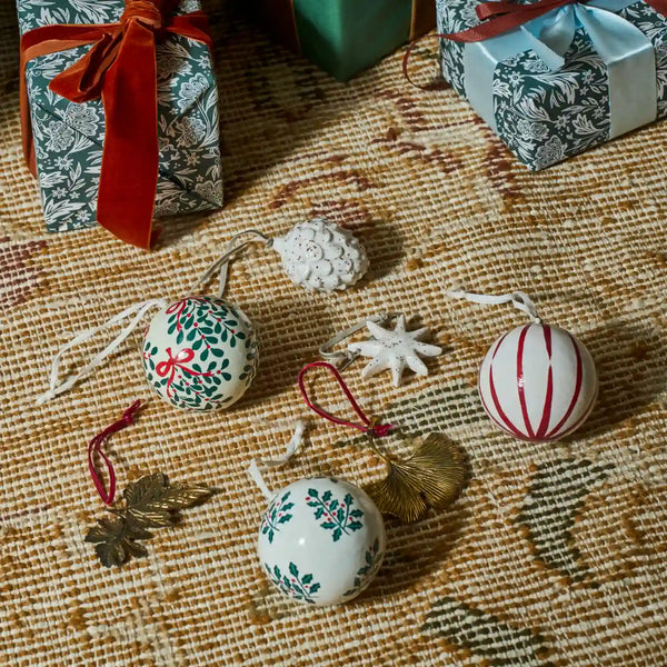 A styled flat-lay of three white ceramic Christmas ornaments with green and red patterns, alongside a white star ornament, a gold leaf ornament, and a small white pinecone, arranged on a textured beige woven rug with wrapped gift boxes in the background.