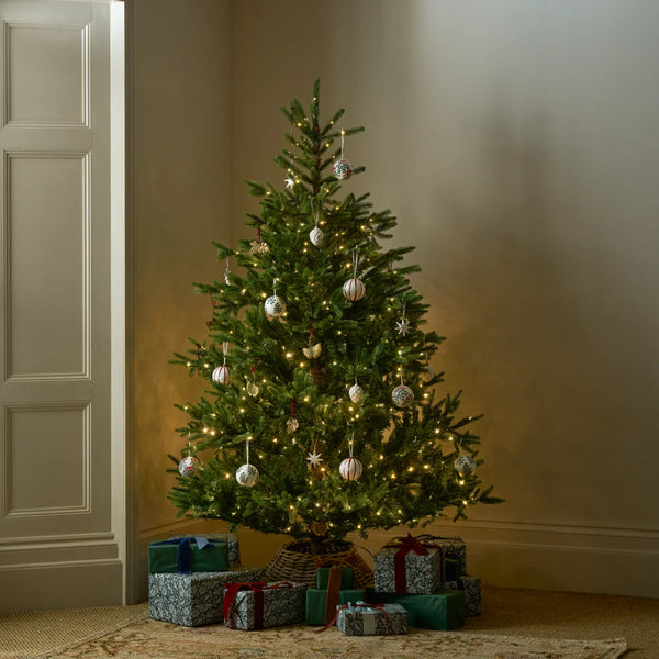 A decorated Christmas tree stands in a corner of a room with wrapped gift boxes underneath. The tree is green with warm white string lights and silver ornaments, set against beige walls and a patterned rug.