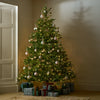A decorated artificial Christmas tree stands in a warmly lit room corner, with wrapped gifts placed at its base. The tree is adorned with warm white string lights and silver, white, and gold ornaments, including baubles, stars, and ribbons.