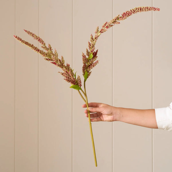 A hand holding a long-stemmed artificial ornamental grass or flax stem with pinkish-brown seed heads and small green leaves, set against a beige paneled wall.