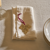 A folded cream linen napkin on a textured tablecloth, decorated with a gold embroidered leaf ornament and a dark red hanging loop.