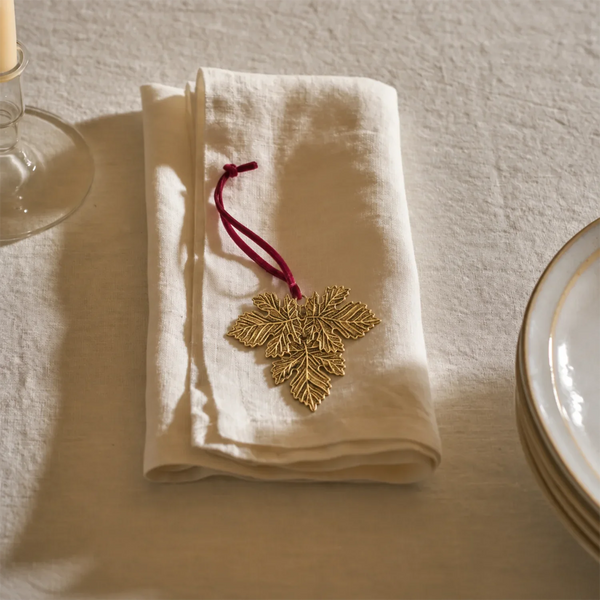 A folded cream linen napkin on a textured tablecloth, decorated with a gold embroidered leaf ornament and a dark red hanging loop.
