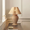 A woven rattan table lamp with a tapered shade and a rounded textured base, lit with a warm glow, set on a dark wooden table in a softly lit room.