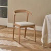 A light natural wood dining chair with a curved backrest and a light beige upholstered seat, shown in a styled room with a wood herringbone floor and a draped tablecloth.