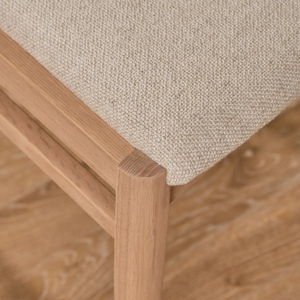 Close-up of a wooden chair corner with a light beige woven fabric seat cushion on a warm wood floor.