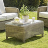 A rectangular wicker coffee table with a glass top sits on a green lawn, styled with a white tray, teapot, cups, and a potted plant. Partial views of matching wicker armchairs with white cushions are visible on both sides.