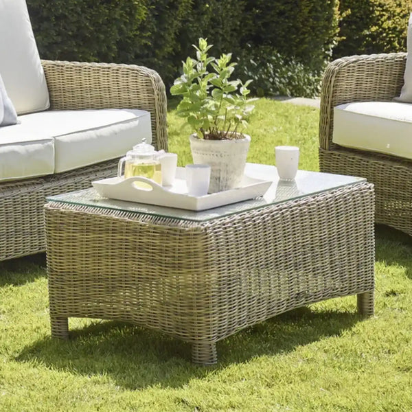 A rectangular wicker coffee table with a glass top sits on a green lawn, styled with a white tray, teapot, cups, and a potted plant. Partial views of matching wicker armchairs with white cushions are visible on both sides.
