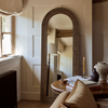 An arched floor mirror with a rustic dark wood frame stands in a cozy, sunlit room with cream paneled walls, a window, and a side table with books and vases.