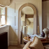 An arched full-length floor mirror with a light natural wood frame, leaning against a cream paneled wall in a cozy, sunlit room.