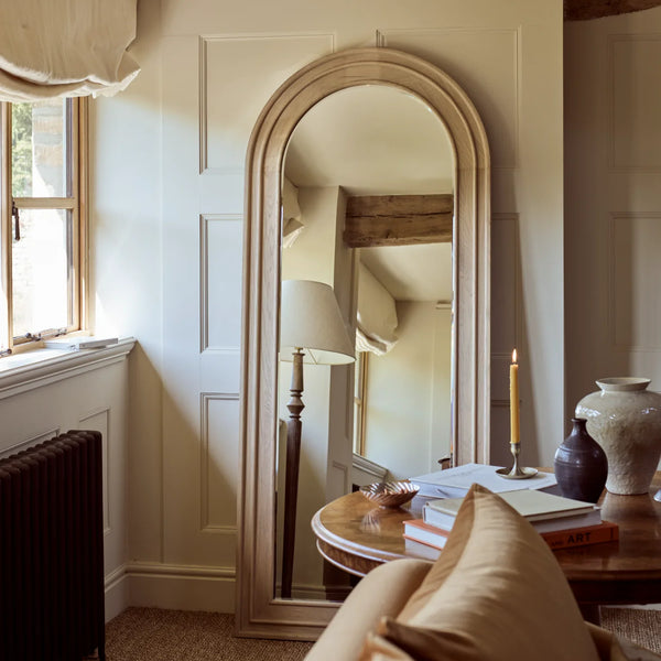 An arched full-length floor mirror with a light natural wood frame, leaning against a cream paneled wall in a cozy, sunlit room.