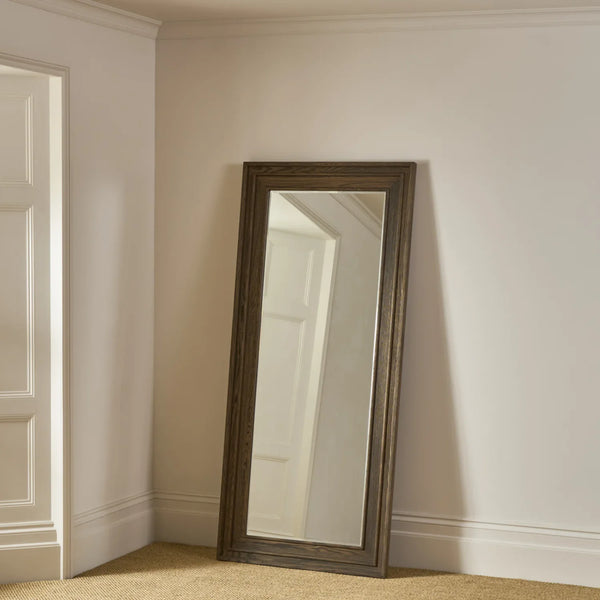 A tall, full-length floor mirror with a distressed dark brown wooden frame, leaning against a beige wall in a neutral room corner.