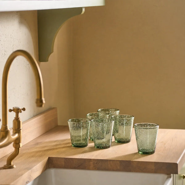 Six green-tinted glass tumblers with a textured, crackled finish arranged on a wooden kitchen counter next to a brass faucet.