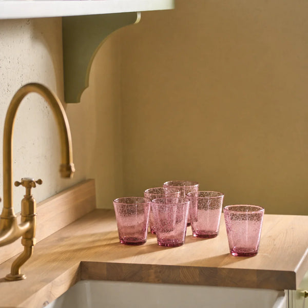 Six pink textured glass tumblers arranged on a wooden kitchen countertop near a brass faucet and sink.