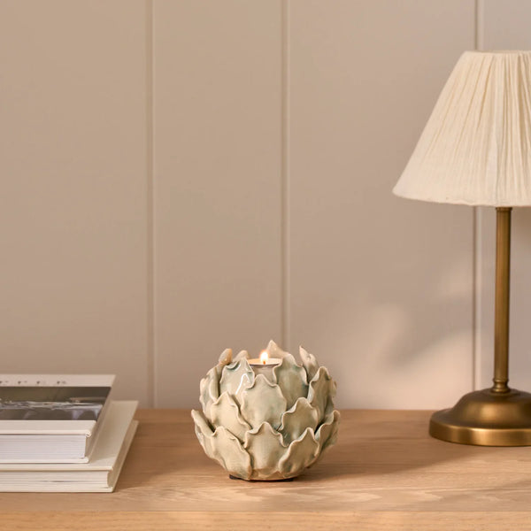 A ceramic artichoke-shaped candle holder with a lit tealight candle, styled on a wooden table next to a lamp and books against a beige paneled wall.