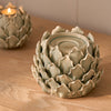 A ceramic artichoke-shaped candle holder with a small tealight candle inside, displayed on a light wood tabletop with another similar candle holder blurred in the background.