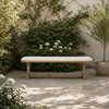 A rectangular outdoor bench with a light beige cushion and natural wood frame, set on a stone patio in a garden with green foliage and white flowers.