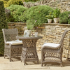 A three-piece outdoor wicker furniture set featuring a round glass-top table and two matching armchairs with white cushions, arranged on a stone patio in front of a rustic stone wall and greenery.