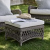 A square outdoor wicker ottoman with a cream cushion sits on a green lawn, styled with an open book and straw hat. Matching patio chairs are partially visible in the background.