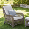 An outdoor wicker armchair with light beige cushions and a blue-gray accent pillow, placed on a green lawn with a stone wall and foliage in the background.