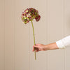 A single artificial-looking hydrangea stem with clustered dusty pink and green petals, held by a person's hand against a beige paneled wall.