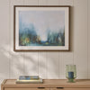 A framed abstract landscape painting with soft blue, green, and gray tones, displayed on a light wood console table against a beige paneled wall. A green glass candle holder and books sit on the table below.