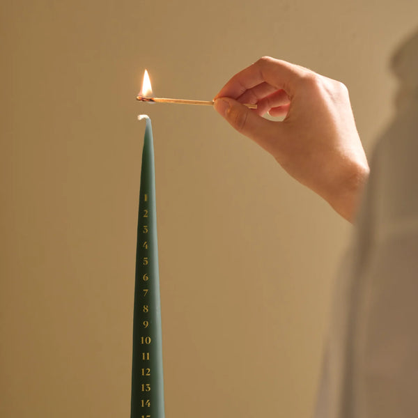 A hand holds a lit match above a tall, slender teal candle marked with numbers from 1 to 15, against a warm beige background.