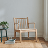 A solid oak Wycombe armchair with a woven rush seat, placed in a room with a neutral color scheme, accompanied by a small wooden side table with a plant and a pair of glasses on top, and some books on the floor.