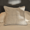 A square decorative throw pillow with a small repeating navy floral block print on an off-white fabric, finished with a matching navy piped border, resting on a bed with white pillows and a dark headboard.