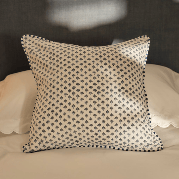 A square decorative throw pillow with a small repeating navy floral block print on an off-white fabric, finished with a matching navy piped border, resting on a bed with white pillows and a dark headboard.