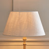 A table lamp with a large tapered drum shade made of textured cream linen fabric, mounted on a light natural wood pedestal base against a neutral beige wall.