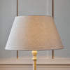 A table lamp with a tapered beige fabric shade and a light wood pedestal base, shown against a softly paneled gray wall.