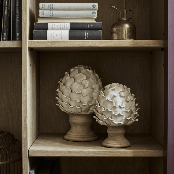 Two decorative ceramic artichokes in cream and beige tones displayed on a wooden bookshelf alongside books and a brass oil can.