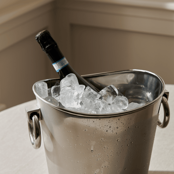 Close-up of the Lydford stainless steel wine bucket with ring handles, filled with ice and a bottle of sparkling wine, with condensation droplets on the polished metal surface.