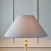 A table lamp with a large conical fabric shade in a light gray color and a light wood base, set against a pale gray paneled wall.