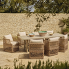 Outdoor dining set with a rectangular light wood table and five woven rattan armchairs with cream cushions, arranged on a paved patio in front of a stone wall.