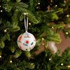 A round white Christmas ornament printed with colorful skiers, hanging from a green Christmas tree with warm string lights in the background.