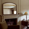 An arched mirror with a dark wood frame hanging above a fireplace mantel in a warm, classic room setting with a lit table lamp and a beige armchair.