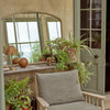 A cozy indoor patio corner featuring a light wood armchair with a textured taupe-gray cushion, surrounded by potted green plants and a large arched mirror reflecting a sunlit window.