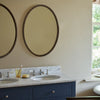 A double bathroom vanity with two undermount sinks, a white marble countertop, and dark navy blue cabinets. Two large oval mirrors with dark wood frames hang on the beige wall above the vanity.