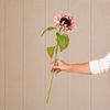 A single artificial-looking pink sunflower with a dark brown center, green stem, and leaves, held by a person's hand against a beige paneled background.