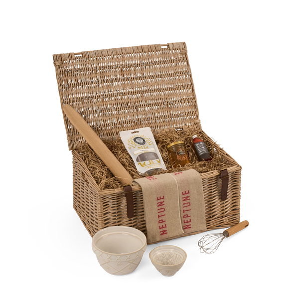 A wicker picnic basket with its lid open, filled with straw padding and food items including jars, a package, and a wooden paddle. In front of the basket are two ceramic bowls, a small dish, and a whisk on a white background.