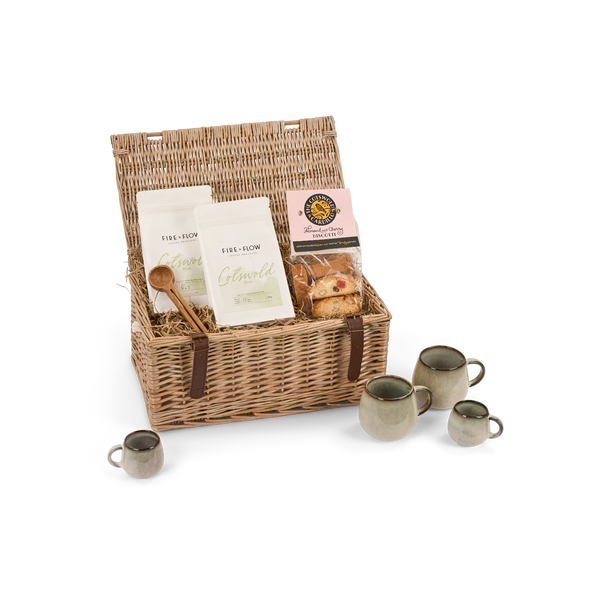 A wicker picnic basket gift set on a white background, containing tea and coffee bags, a small pastry, a wooden spoon, and four ceramic mugs.
