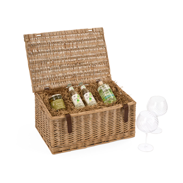 A natural wicker picnic hamper with its lid open, containing a bottle of craft gin and 2 bottles of premium tonic water with a jar of preserves nestled in shredded paper, with two clear stemmed gin glasses beside it