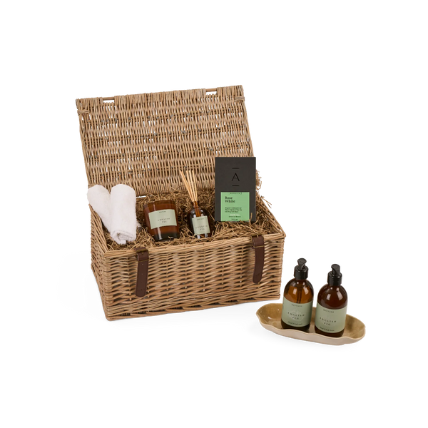 A wicker gift basket with an open lid, filled with spa items including a white towel, amber bottles, a reed diffuser, and a soap bar, shown on a white background with two small bottles on a tray in front.