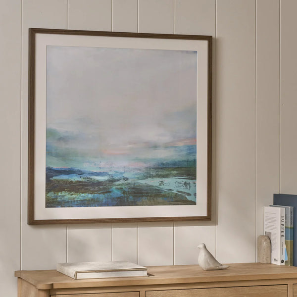 A framed abstract landscape art print with a brown frame and white mat, hanging on a beige wood-paneled wall above a light wood console table with books and a small bird figurine.