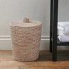 A natural woven rattan bin with a matching lid and top loop, set on a wooden floor next to a black shelving unit with white towels.