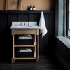An undermount open bathroom washstand with turned column legs, featuring a white Carrara marble top and oak wood frame and shelving, in a room with dark walls and a window.